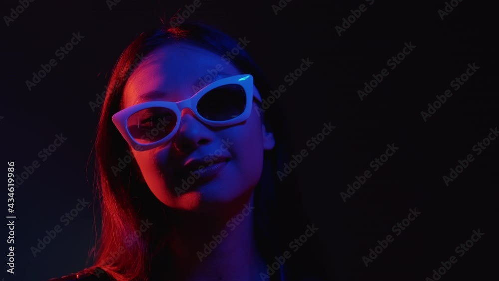 Neon light face. Clubber lifestyle. Synthwave party. Disco night. Blue red color relaxed Asian girl in stylish glasses enjoying rhythm dancing on dark copy space background. Neon light face. Clubber lifestyle. Synthwave party. Disco night. Blue red color relaxed Asian girl in stylish glasses enjoying rhythm dancing on dark copy space background.