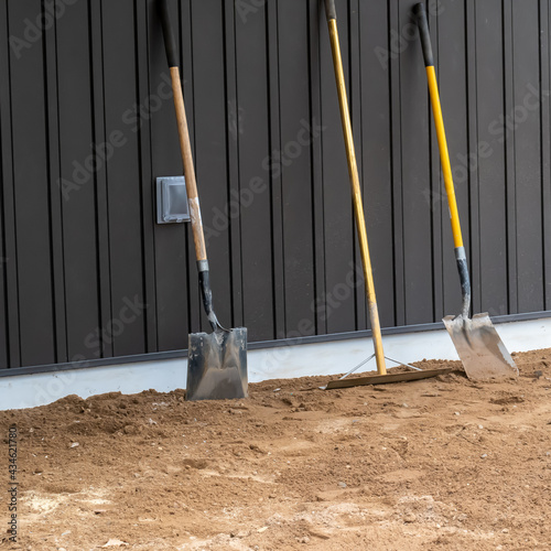 Dirty, used shovels and a rake stand upright on the dirt and lean against the board and batton siding of a wall at a construction site. photo