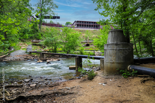 a shot of the rushing water of Big Creek river with a pipe across the river and buildings on a hillside covered with lush green trees and plants at Vickery Creek in Roswell Georgia photo