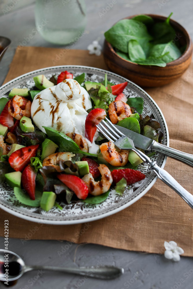 Shrimp, spinach, strawberry and burrata salad in a large vintage plate on a gray table. Healthy food with vegetables, cheese and seafood. Flat lay.