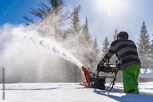 Rear view of a young man pushing an orange mechanical snowplough in the knee deep fresh snow, with a big jet of snow exiting the machine.