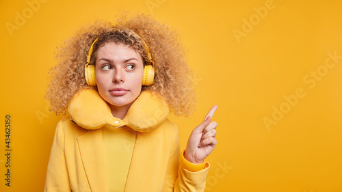 Studio shot of serious curly haired millennial female model gives recommendation indicates at blank copy space wears stereo headphones on ears travel pillow around neck isolated on yellow wall © wayhome.studio 