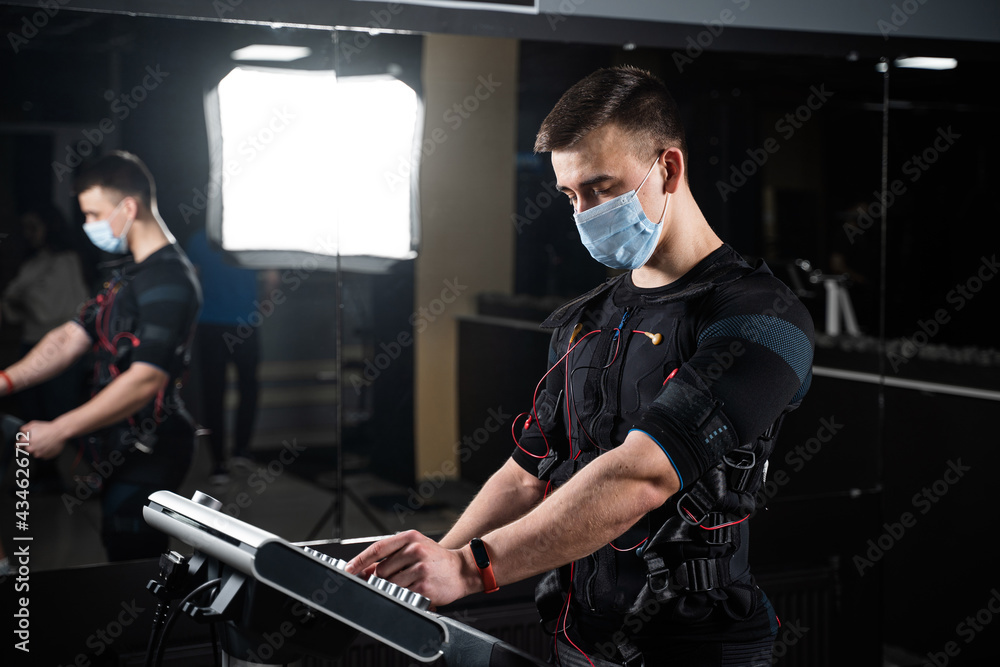 Man in EMS suit and medical mask in gym. Protection from coronavirus covid-19. Sport training in electrical muscle stimulation suit at quarantine period.
