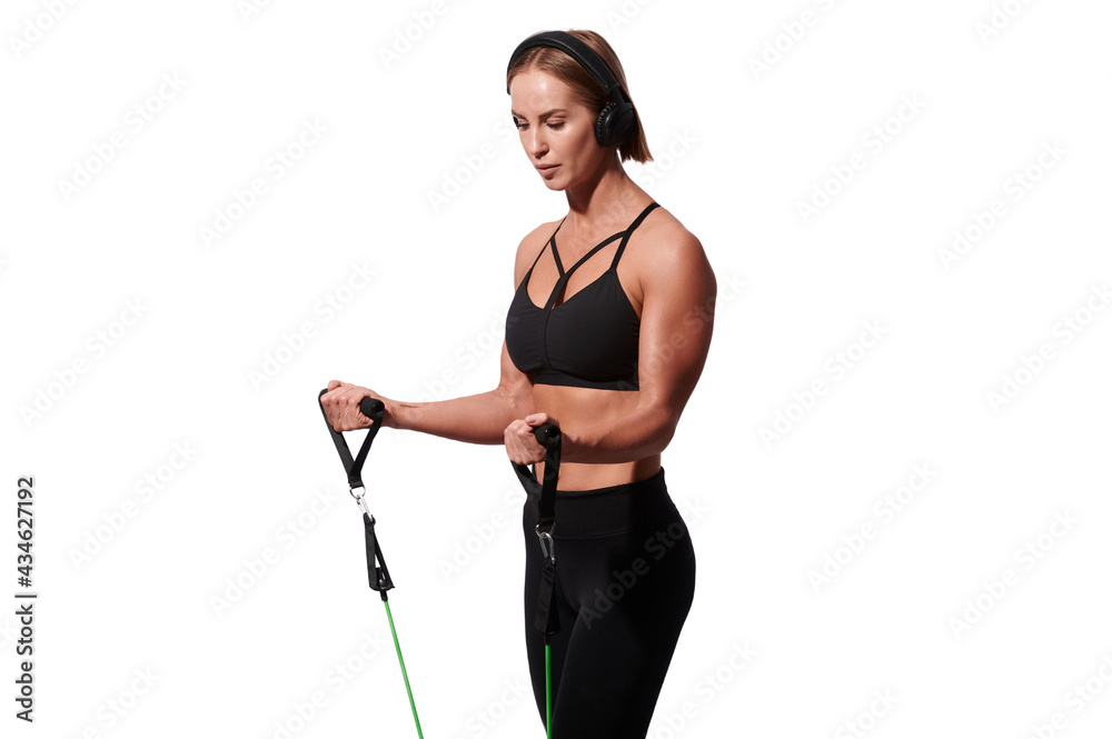 Strong muscular sportswoman in headphones exercising with resistance band over white isolated background