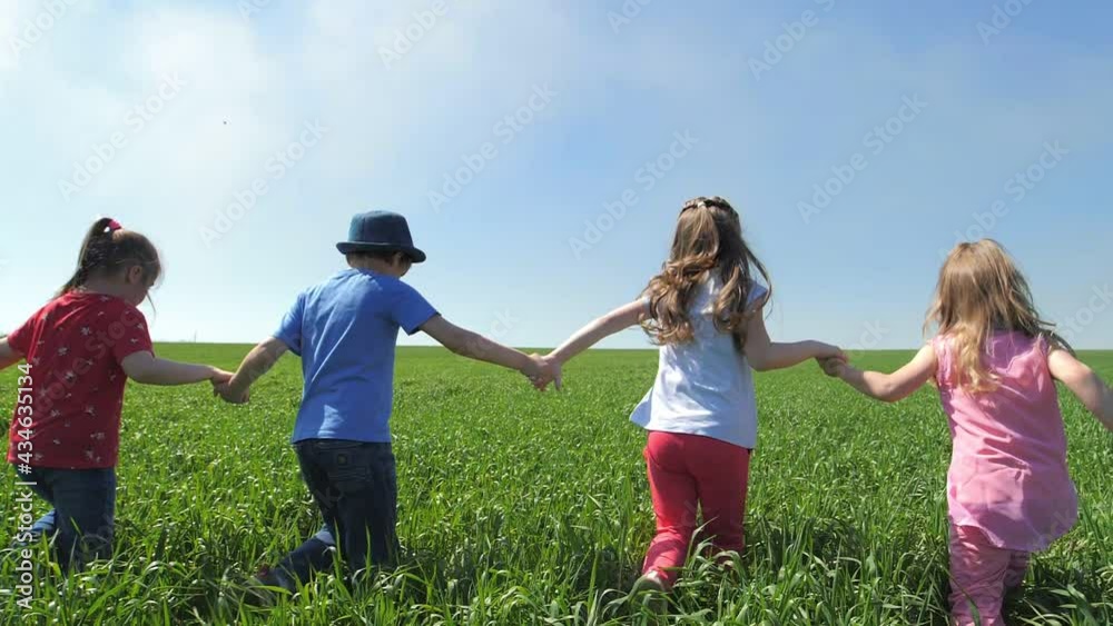 Happy children, boys and girls, holding hands, ran merrily in the fresh air on a green field. Children play catch-up in the park. The concept of children's happiness. Back view . Slow motion Happy children, boys and girls, holding hands, ran merrily in the fresh air on a green field. Children play catch-up in the park. The concept of children's happiness. Back view . Slow motion