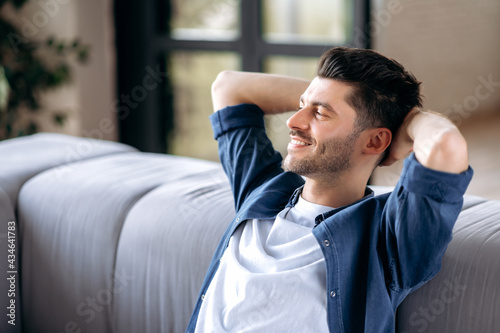 Closeup of happy bearded caucasian adult man in casual clothes relaxing on couch at home. Relaxed guy having break on sofa in living room, dreaming about vacation, looks away, smiling.