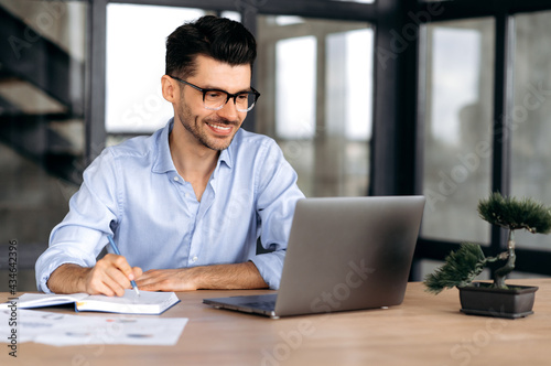 Online education. Intelligent successful caucasian man with glasses, sitting at the workplace, using a laptop, takes notes, studying or working online, listening to a webinar, gaining knowledge, smile