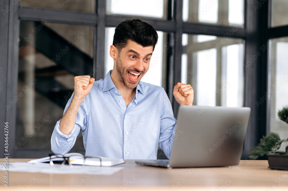 Happy excited caucasian male freelancer or office worker, sitting at his work desk at office, using laptop, shouts and rejoice in success, win or good profit, looking at laptop and gesturing hands Happy excited caucasian male freelancer or office worker, sitting at his work desk at office, using laptop, shouts and rejoice in success, win or good profit, looking at laptop and gesturing hands