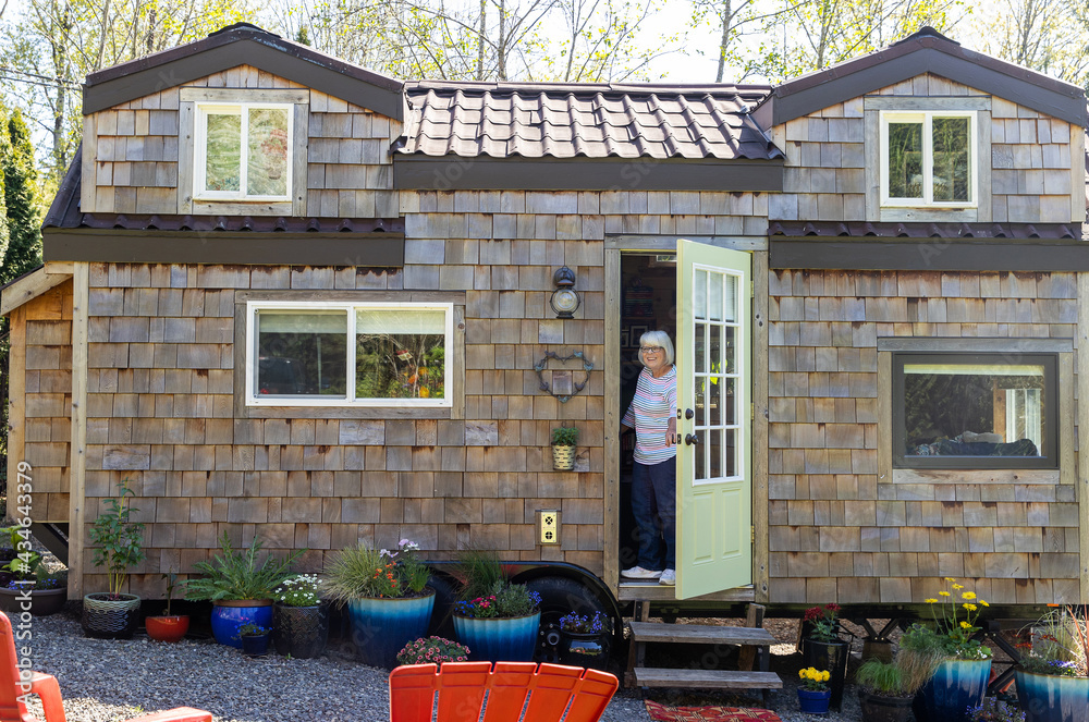 Friendly mature woman standing at front door of her charming little wooden tiny home or house - obrazy, fototapety, plakaty 