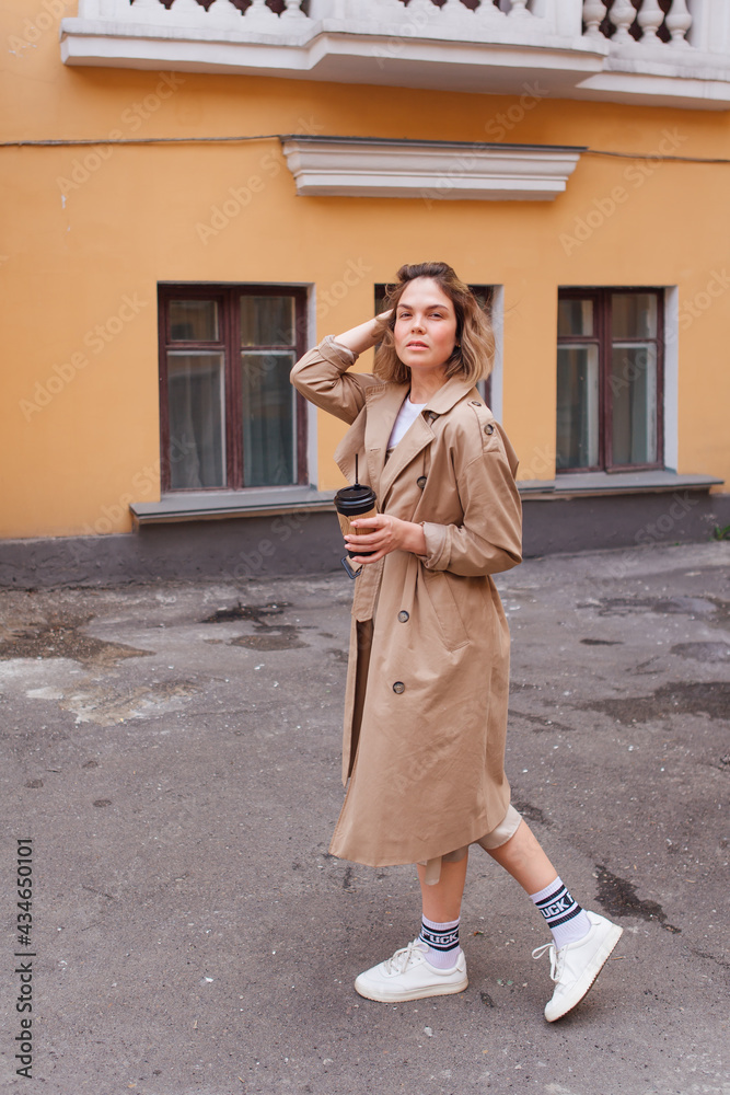 Young millennial woman with wild hair dressed in an autumn coat walking with a cup of coffee to go near the old building.