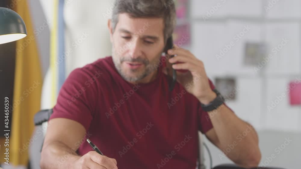 Mature Male Fashion Designer In Wheelchair In Studio Working On Sketches And Talking On Mobile Phone Mature Male Fashion Designer In Wheelchair In Studio Working On Sketches And Talking On Mobile Phone
