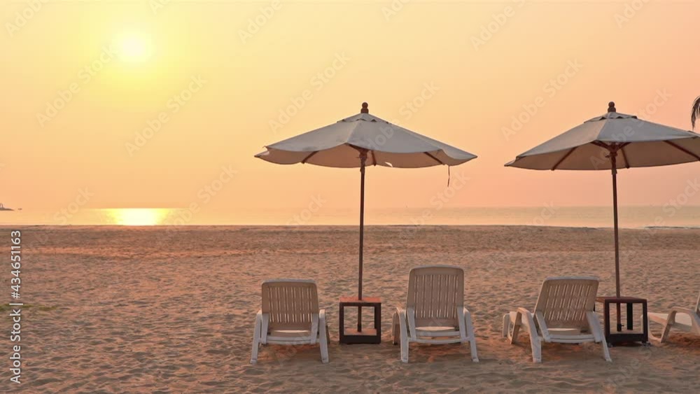 A couple of plastic seats and umbrellas on an empty white sand beach with golden sun over the ocean during sunset.