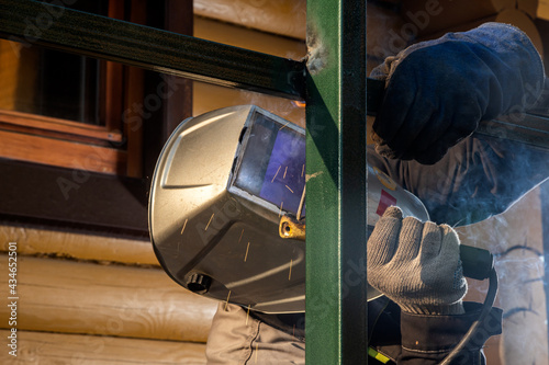 Close up of a young  man welder in  uniform, welding mask and welders leathers, weld  metal  with a  welding machine at the construction  of a fence in summer day putside in the village photo