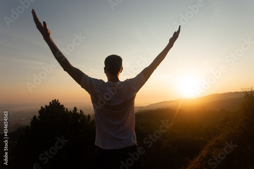Silhouette of young man raises his hands up to sunset or sunrise on on top of hill in the summer 
