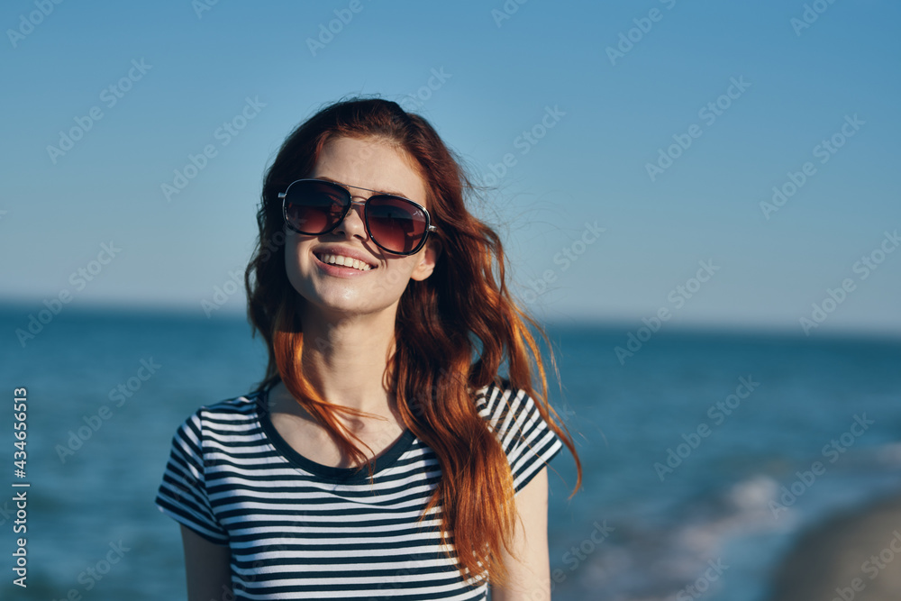 woman in sunglasses and a striped t-shirt gestures with her hands in the mountains near the sea on the beach