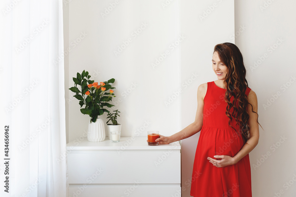 Young beautiful pregnant woman, dressed in a red dress, standing at home near the chest of drawers and drinking juice.