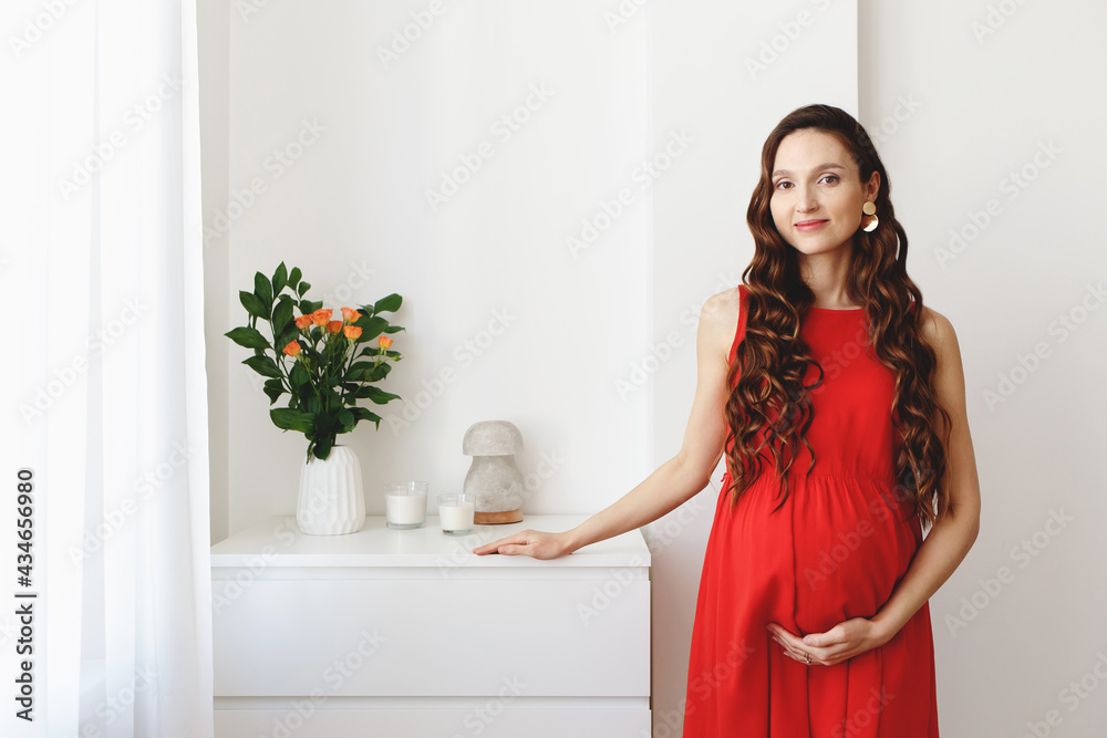 Young beautiful pregnant woman, dressed in a red dress, standing at home near the chest of drawers. Young beautiful pregnant woman, dressed in a red dress, standing at home near the chest of drawers.