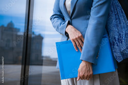 A business woman in a blue jacket holds a folder with papers in her hand in front of a glass office building. Concept of successful women
