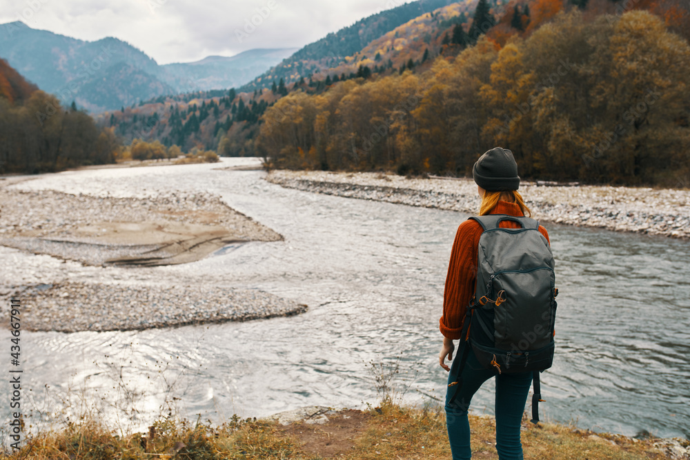 a woman in a red jacket and a backpack on her back are walking on the banks of the river in the mountains
