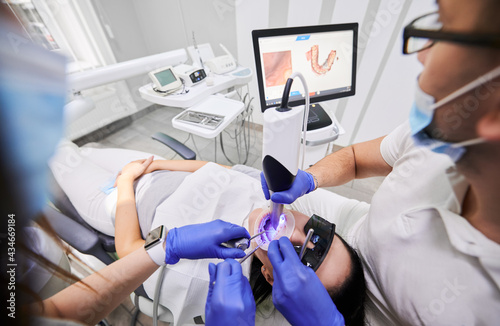 Close up of dentist and assistant using dental intraoral scanner while examining patient teeth in dental office. Specialist in sterile gloves performing intraoral scanning with modern scanning machine