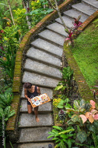 Woman carrying breakfast tray down curved stair case through jungle forest to a bungalow in Bali.
