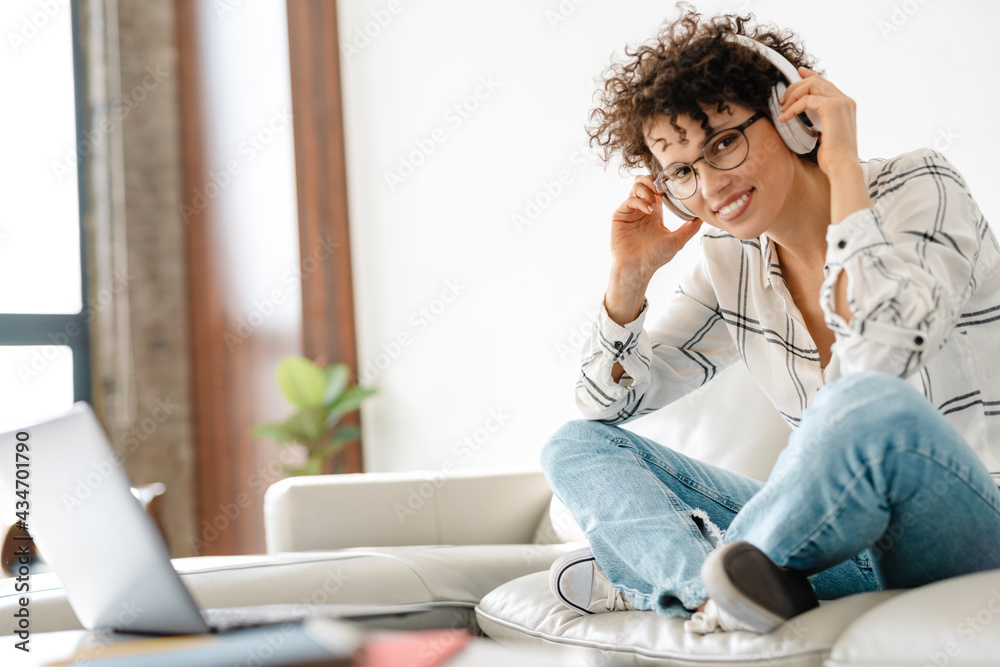 Young curly woman in headphones using laptop while sitting on couch Young curly woman in headphones using laptop while sitting on couch