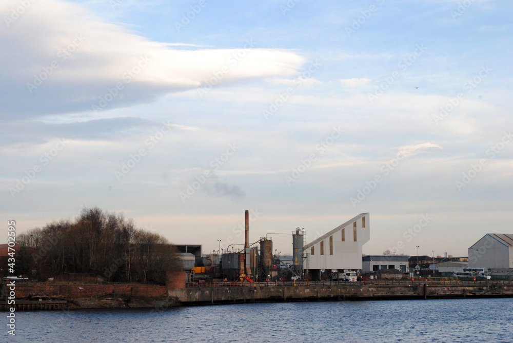 Chimney & Industrial Buildings seen across River against Cloudy Sky 