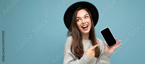 Panoramic Closeup of charming young happy woman wearing black hat and grey sweater holding phone looking to the side pointing finger at screen isolated on background photo