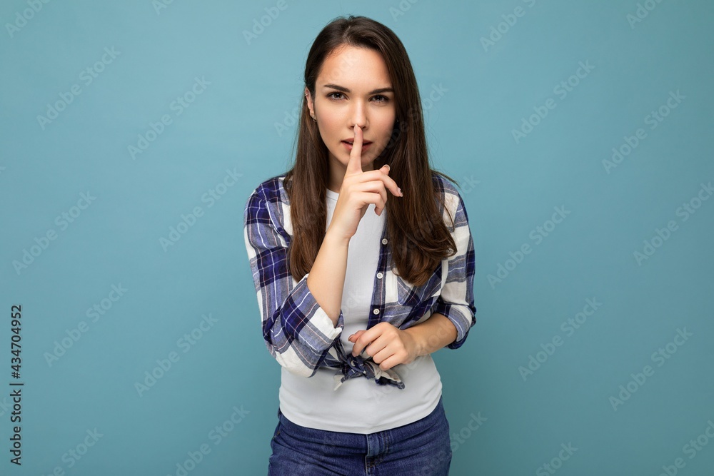 Young serious beautiful brunet woman with sincere emotions wearing trendy check shirt standing isolated on blue background with empty space and showing shhh gesture. Keep secret concept Young serious beautiful brunet woman with sincere emotions wearing trendy check shirt standing isolated on blue background with empty space and showing shhh gesture. Keep secret concept