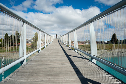 A man walking on the bridge of Lake Tekapo, with bridge railing casting a long shadow, South Island