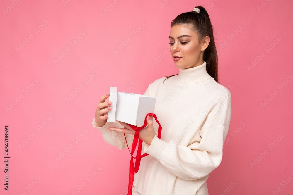 Attractive happy amazed young brunette woman isolated over pink background wall wearing white sweater holding gift box and unboxing present looking at camera Attractive happy amazed young brunette woman isolated over pink background wall wearing white sweater holding gift box and unboxing present looking at camera
