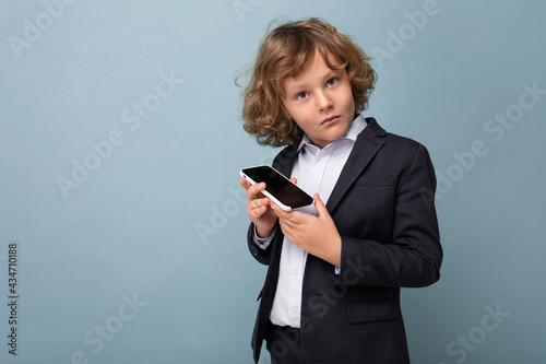 Handsome boy with curly hair wearing suit holding phone isolated over blue background looking at camera photo