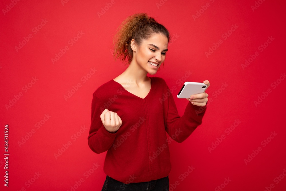 Young emotional happy woman wearing dark red sweater isolated over red background holding smartphone having fun and showing yes gesture looking at mobile phone screen Young emotional happy woman wearing dark red sweater isolated over red background holding smartphone having fun and showing yes gesture looking at mobile phone screen