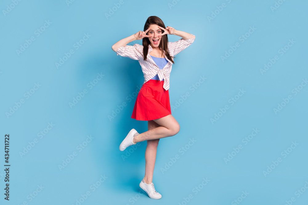 Full size portrait of charming cheerful lady stand on one foot fingers show v-sign near eyes isolated on blue color background Full size portrait of charming cheerful lady stand on one foot fingers show v-sign near eyes isolated on blue color background