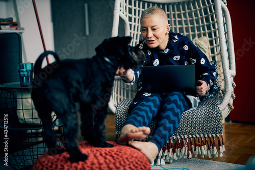 Smiling cute senior woman with short hair sitting in her chair with laptop in her lap and playing with her dog.