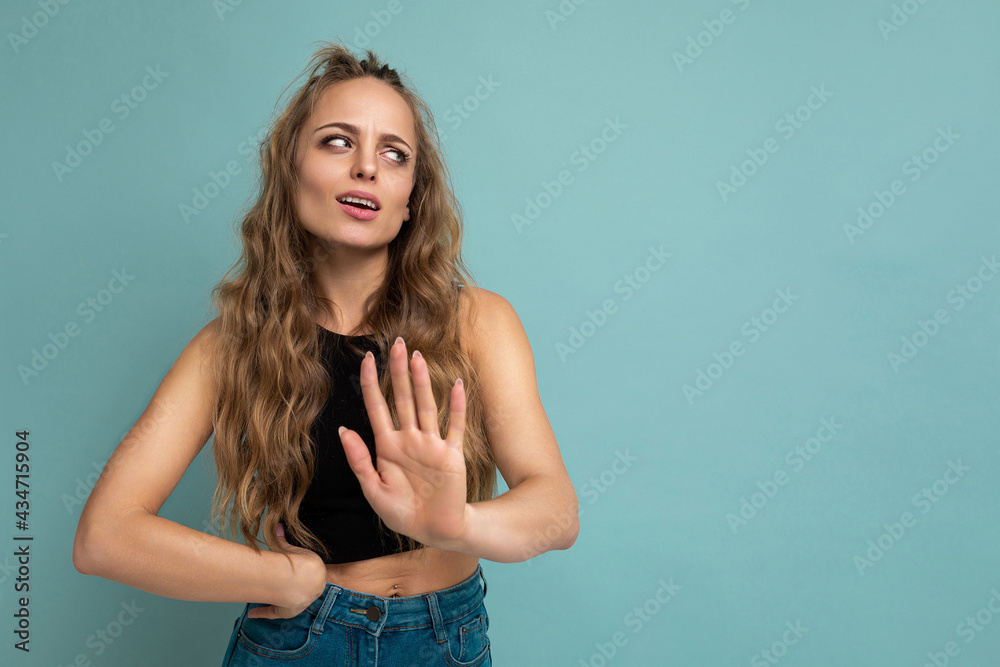 Young tired upset attractive winsome blonde curly woman with sincere emotions wearing trendy black top isolated over blue background with free space and showing stop gesture saying no. Negative