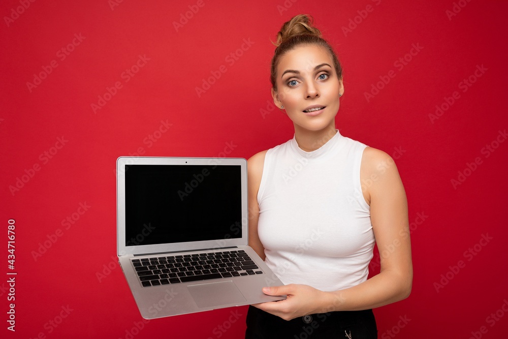 Naklejka premium beautiful blond young woman looking at camera holding computer laptop wearing white t-shirt isolated over red wall background