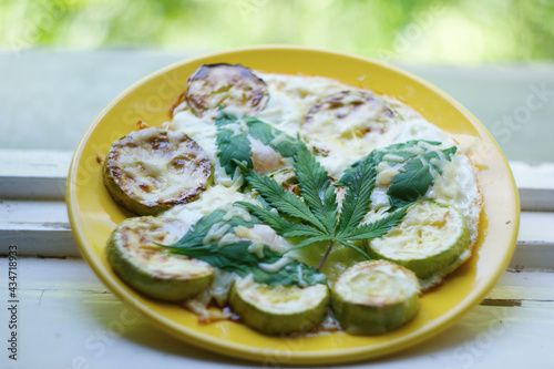 cannabis food. classic cannabis leaf lies on a fried egg baked with zucchini and marijuana leaves. grated cheese sprinkled on top. a plate of food lies on an old rustic window overlooking the garden