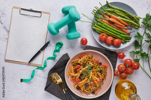 Diet plan, menu or program, tape measure, water, dumbbells and diet food, top view. Vegetarian vegetable pasta. Tasty appetizing classic italian spaghetti pasta with tomato sauce. salad. clipboard photo