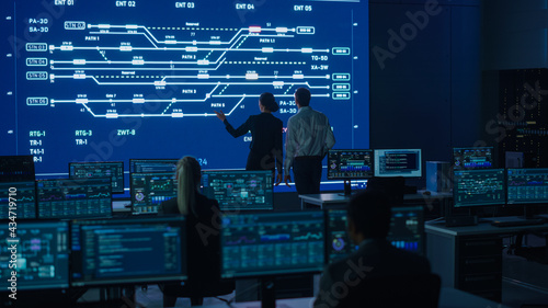 Project Manager and Computer Science Engineer Talking while Using Big Screen Display Showing Infrastructure Infographics and Data.Telecommunications Control Monitoring Room with People Working