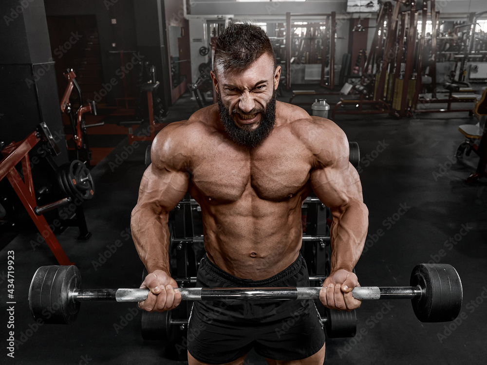 A brutal man with a beard in the gym. A muscular bodybuilder in perfect athletic shape performs a bicep exercise, lifting a barbell. Emotional training. Concept of sports and a healthy lifestyle A brutal man with a beard in the gym. A muscular bodybuilder in perfect athletic shape performs a bicep exercise, lifting a barbell. Emotional training. Concept of sports and a healthy lifestyle