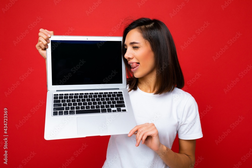 Photo shot of funny smiling Beautiful happy overjoyed young woman with short brunet hair crazy sexy tongue and close eyes holding computer laptop wearing white t-shirt and jeans isolated over red wall Photo shot of funny smiling Beautiful happy overjoyed young woman with short brunet hair crazy sexy tongue and close eyes holding computer laptop wearing white t-shirt and jeans isolated over red wall
