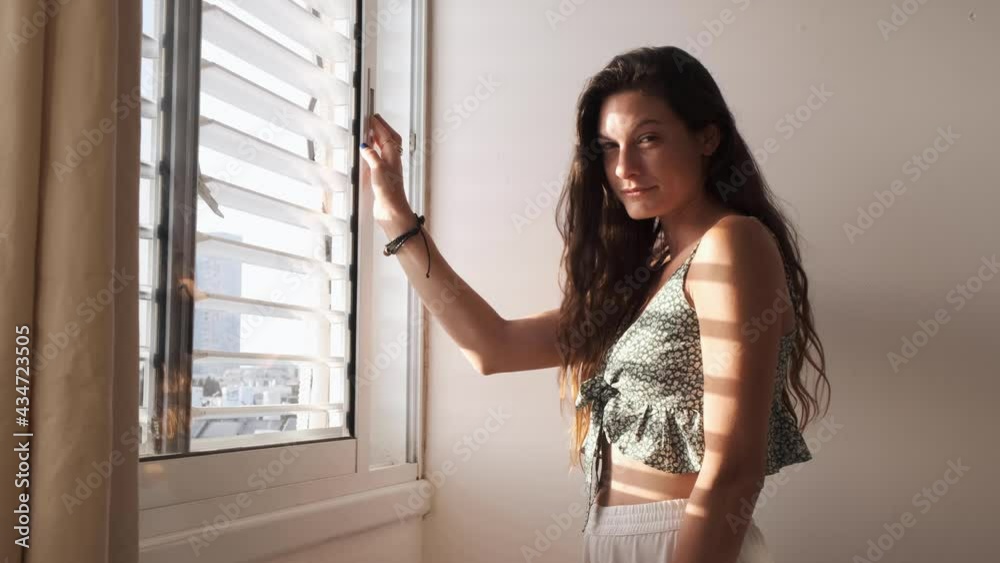 Woman opening the window and letting the sun shine on her body. MEDIUM shot. a beautiful lady standing in front of the window looking outside with hope. sunlight shining on her body. morning sun Woman opening the window and letting the sun shine on her body. MEDIUM shot. a beautiful lady standing in front of the window looking outside with hope. sunlight shining on her body. morning sun