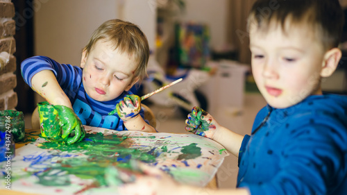 Two boys are painting at the table at home, smaller is in focus, another is blurred. Hands are smudged with paint and painting is messy. photo