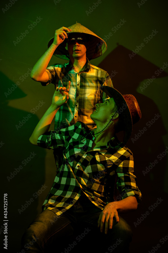 Two guys in cowboy and Chinese hats on a dark background illuminated by yellow and green light.