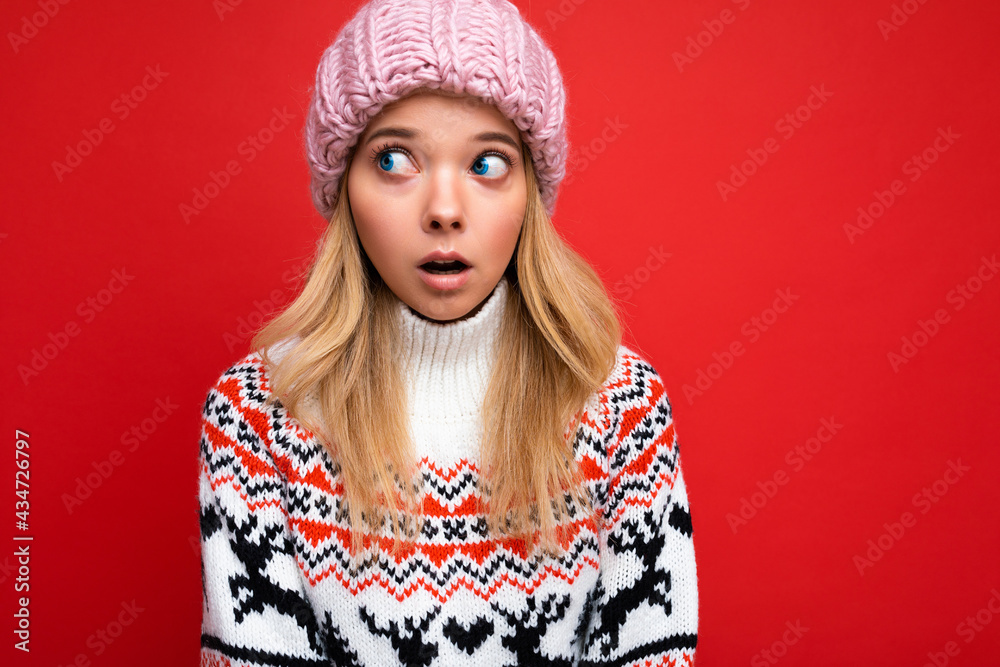 Photo of beautiful shocked young blonde woman isolated over red background wall wearing winter sweater and pink hat looking to the side Photo of beautiful shocked young blonde woman isolated over red background wall wearing winter sweater and pink hat looking to the side