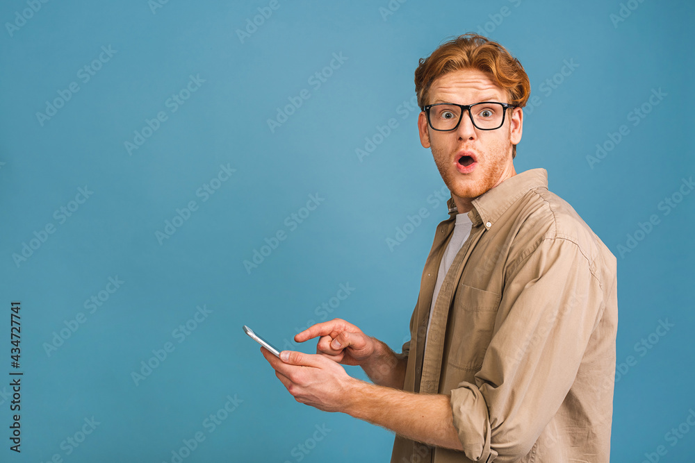 Portrait of young amazed shocked man in casual typing sms isolated over blue background. Using phone. Portrait of young amazed shocked man in casual typing sms isolated over blue background. Using phone.