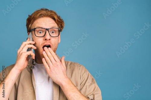 Fototapeta Naklejka Na Ścianę i Meble -  Oh, great news! Smiling shocked amazed young casual man talking on the mobile phone isolated over blue background.