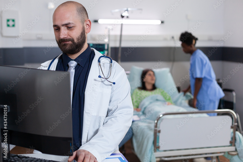 Front view of practitioner doctor typing medical expertise on computer while in background black asisstant discussing healthcare treatment. Hospitalized patient having respiratory disorder Front view of practitioner doctor typing medical expertise on computer while in background black asisstant discussing healthcare treatment. Hospitalized patient having respiratory disorder