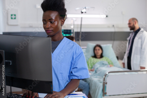 Black medical asisstant typing sickness treatment on computer while physician doctors monitoring sick woman analyzing diagnosis. Patient lying in hospital ward bed during consultation recovery